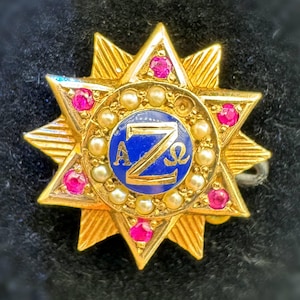 May include: A gold-toned star-shaped pin with a blue center featuring the letters "A Z Ω". The pin is adorned with small pink gemstones and pearls. The pin has a textured, layered design.