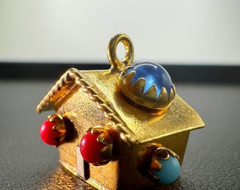 May include: A gold-colored house pendant with a loop for a chain. The roof has a blue gemstone and the sides have red and turquoise gemstones. The pendant is likely made of metal and has a detailed design.