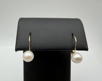 May include: A pair of drop earrings featuring round, white pearls suspended from thin, curved gold-toned metal wires. The earrings are displayed on a black stand, highlighting the contrast between the lustrous white pearls and the metallic gold hooks.
