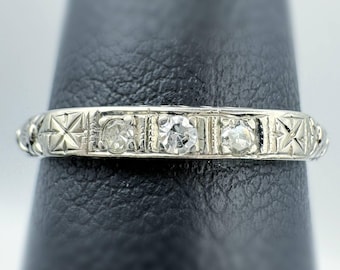 Peut inclure: Une bague en argent ornée de trois pierres précieuses transparentes disposées en rangée. La bande présente un motif géométrique complexe avec des motifs en forme d'étoile. La bague est présentée sur une surface noire.
