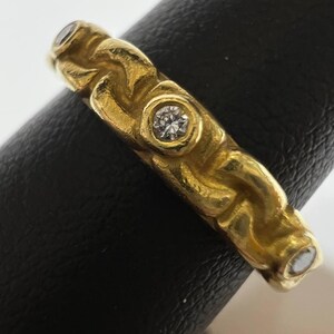 Puede incluir: Un anillo de oro con un dise&ntilde;o texturizado y tres peque&ntilde;as piedras preciosas claras. El anillo se muestra sobre una superficie negra, mostrando sus intrincados detalles y su color c&aacute;lido.