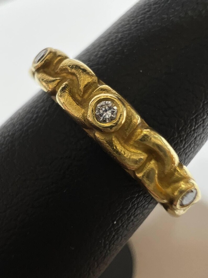 Puede incluir: Un anillo de oro con un dise&ntilde;o texturizado y tres peque&ntilde;as piedras preciosas claras. El anillo se muestra sobre una superficie negra, mostrando sus intrincados detalles y su color c&aacute;lido.