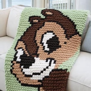 May include: A crocheted blanket featuring a pixelated image of a cartoon chipmunk's face. The blanket uses brown, tan, black, white, and green yarn. The blanket is draped over a white couch.