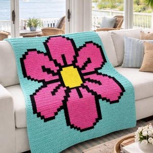 May include: A crocheted blanket featuring a large pink flower with a yellow center and black outlines, set against a light blue background. The blanket is draped over a white sofa, creating a cozy and colorful accent.