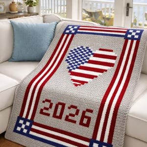 May include: A gray knitted blanket with a heart-shaped American flag design. The blanket features red and white stripes, blue accents, and the year "2026" in red. It is draped over a white sofa.
