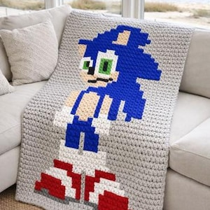 May include: A crocheted blanket featuring a pixelated image of Sonic the Hedgehog. The blanket is mainly grey, with blue, white, red, and green details. The blanket is draped over a white sofa near a window.