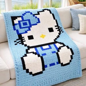 May include: A light blue crocheted blanket featuring a pixelated image of a cartoon cat. The cat has a white face, black eyes and whiskers, a yellow nose, and a blue bow. The blanket is draped over a white sofa.