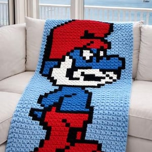 May include: A crocheted blanket featuring a pixelated image of a Smurf character. The blanket is primarily light blue with the Smurf's red hat and boots, blue body, and white face. The blanket is draped over a white couch.