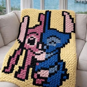 Puede incluir: Una manta amarilla tejida a ganchillo con un dise&ntilde;o pixelado de dos personajes de dibujos animados, uno rosa y otro azul, con contornos negros. La manta est&aacute; sobre un sof&aacute; blanco, con vistas al mar al fondo.
