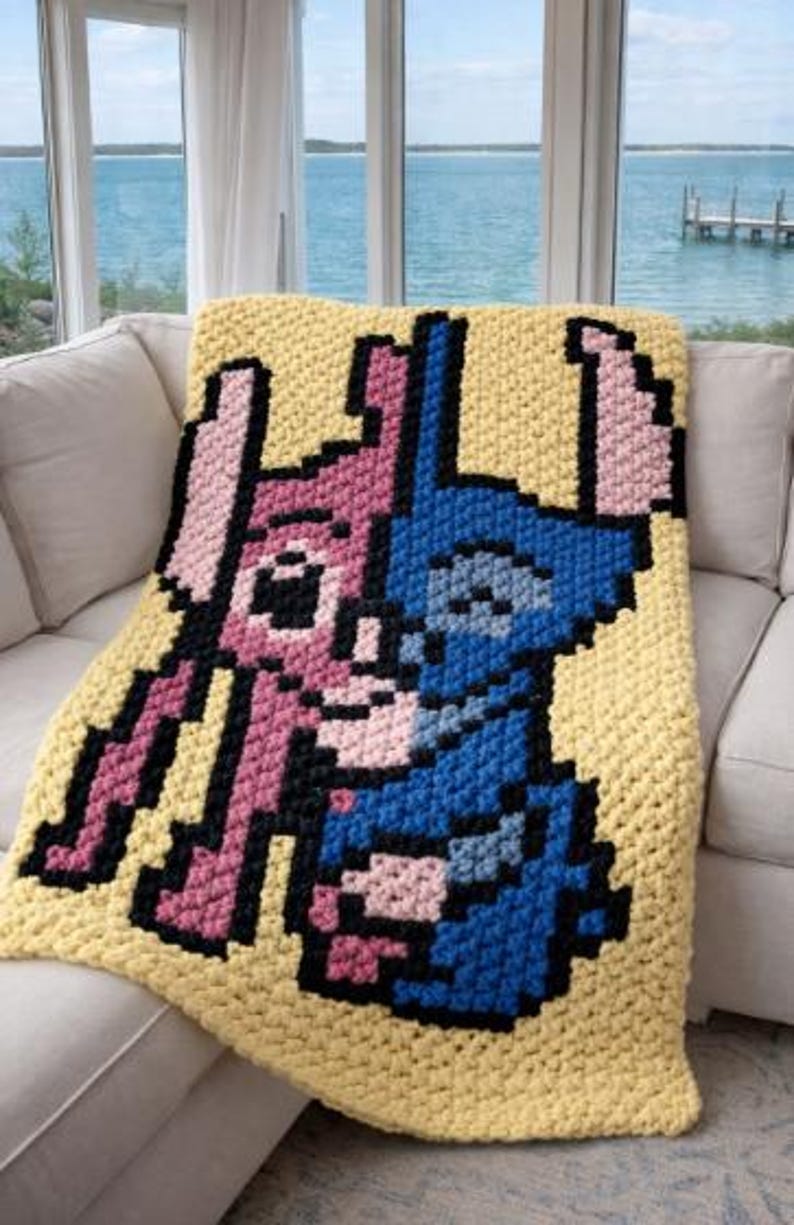 Puede incluir: Una manta amarilla tejida a ganchillo con un dise&ntilde;o pixelado de dos personajes de dibujos animados, uno rosa y otro azul, con contornos negros. La manta est&aacute; sobre un sof&aacute; blanco, con vistas al mar al fondo.