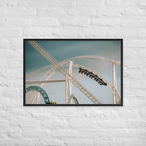 Puede incluir: Impresión enmarcada de una montaña rusa contra un cielo azul. La pista blanca se curva y forma bucles, con un tren de personas en la parte superior de un bucle. Un bucle verde y blanco es visible en el fondo.