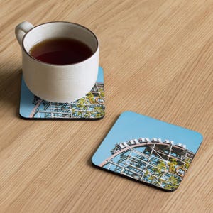 May include: Square coaster set featuring a roller coaster design with a blue sky background. A white ceramic mug filled with tea sits on one coaster. The coasters are on a light wood surface.