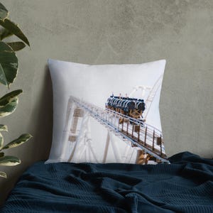 Puede incluir: Cojín cuadrado blanco con una imagen de una montaña rusa. Los vagones son negros y azules, subiendo por una vía blanca. El cielo está nublado. El cojín mide aproximadamente 40 cm x 40 cm.