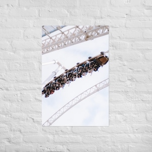 Puede incluir: Impresión de póster que muestra una montaña rusa con pasajeros. El vagón de la montaña rusa está inclinado hacia abajo, lleno de gente. El cielo está nublado y la estructura de soporte de la montaña rusa es visible.