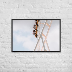 Puede incluir: Impresión enmarcada de una montaña rusa ascendiendo una pendiente pronunciada. Los vagones amarillos y negros están llenos de pasajeros con los brazos levantados. La vía es blanca, sobre un cielo nublado. La impresión está en un marco negro.