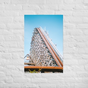 May include: A photograph of a wooden roller coaster against a clear blue sky. The coaster's structure is made of brown wood, with the track visible at the top. The image is a vertical shot, capturing the height and scale of the ride.
