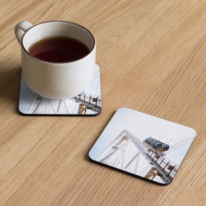 Puede incluir: Una taza de cerámica blanca llena de té descansa sobre un posavasos con una imagen de montaña rusa. Un segundo posavasos con la misma imagen está al lado de la taza. Los posavasos tienen un borde negro.