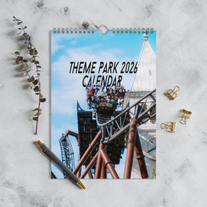 May include: A spiral-bound calendar with the text "THEME PARK 2026 CALENDAR" against a blue sky background. The image shows a roller coaster with people riding it. A pen and gold binder clips are also present.