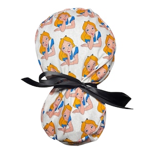 May include: A white fabric scrub cap featuring a repeating pattern of Alice from Alice in Wonderland. Alice is depicted with blonde hair and a blue dress. The cap is tied with a black ribbon.