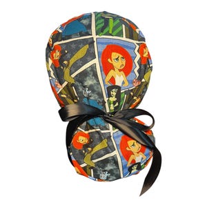 May include: A scrub cap featuring a comic book-style print with red-haired characters in action poses. The cap is tied with a black satin ribbon. The background is a mix of blue, green, and gray tones.