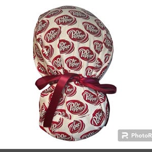 May include: A white and red patterned scrub cap featuring the Dr. Pepper logo. The cap is tied with a burgundy ribbon. The fabric is covered in repeating Dr. Pepper logos.