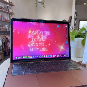 May include: A rose gold MacBook Air laptop with a pink and white desktop background that says "Lay the house down Boots Kitty Girl".