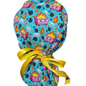 May include: A teal scrub cap featuring a repeating pattern of pink dumpsters on fire with smiling faces, orange flames, black smoke clouds, and the text THIS IS FINE. The cap is secured at the back with a bright yellow satin ribbon tie.