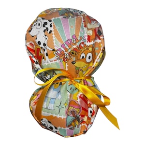 May include: A colorful scrub cap featuring a repeating pattern of cartoon characters, including a Dalmatian, a fox, and a blue dog. The cap is tied with a bright yellow ribbon. The words "Girl Gang" are printed on the cap.