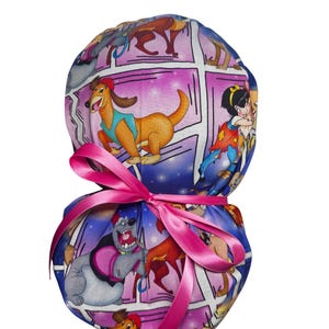 May include: A patterned scrub cap featuring cartoon dog characters in a comic book style. The cap is tied with a bright pink ribbon. The background has a purple and blue color scheme.