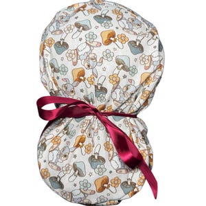 May include: A white patterned scrub cap featuring bunnies, mushrooms, and flowers in pastel colors. The cap is tied with a burgundy ribbon. The fabric design includes light blue, yellow, and brown elements.