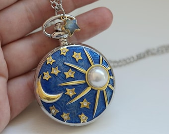 May include: A celestial-themed pendant necklace featuring a round, blue enamel pocket watch design. The watch face has a sun, moon, and star motif with gold accents and a pearl center. The pendant hangs from a silver chain.