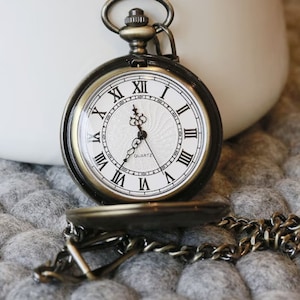 K&ouml;nnte beinhalten: Antike bronzefarbene Taschenuhr mit wei&szlig;em Zifferblatt und schwarzen r&ouml;mischen Ziffern. Die Uhr hat eine Kette und ist teilweise ge&ouml;ffnet, wodurch die Zeit sichtbar wird. Das Wort "QUARTZ" ist auf dem Zifferblatt aufgedruckt.
