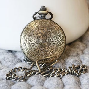 K&ouml;nnte beinhalten: Antike Taschenuhr aus Messing mit einem verzierten Blumendesign auf der Vorderseite. Die Uhr hat eine passende Kette. Sie liegt auf einer grauen, strukturierten Oberfl&auml;che. Oben befindet sich eine kleine Schlaufe.