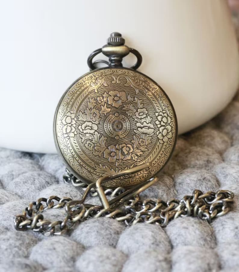 K&ouml;nnte beinhalten: Antike Taschenuhr aus Messing mit einem verzierten Blumendesign auf der Vorderseite. Die Uhr hat eine passende Kette. Sie liegt auf einer grauen, strukturierten Oberfl&auml;che. Oben befindet sich eine kleine Schlaufe.