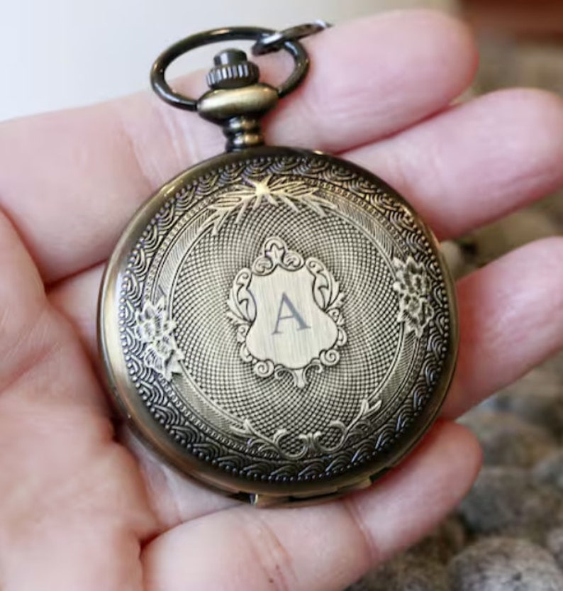 K&ouml;nnte beinhalten: Antike bronzefarbene Taschenuhr mit aufwendigen floralen und scrollartigen Details. Das Zifferblatt der Uhr zeigt ein Schilddesign mit dem Buchstaben "A" in der Mitte. Sie hat eine Schlaufe f&uuml;r eine Kette und eine Aufzugswelle.
