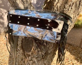 May include: Clear plastic clutch bag with a black and white cowhide panel and silver studs. The bag has black leather fringe on the bottom.