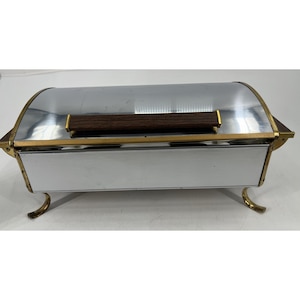 May include: A rectangular chrome bread box with a curved, roll-top lid featuring a dark brown wood-grain handle. The container is accented with gold-toned metal trim along the edges and is supported by four curved, gold-toned metal legs.