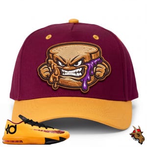 May include: A maroon and gold baseball cap with an embroidered cartoon peanut butter and jelly sandwich with an angry expression. The cap has a yellow brim and a yellow button on top. A yellow and black trainer is in the foreground.