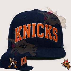 May include: A navy blue flat-brimmed baseball cap with the word "KNICKS" in orange block letters. The cap features a green underside and a small graphic of a basketball player holding a trophy. The cap is a sports-themed accessory.
