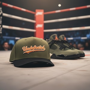 Könnte beinhalten: Eine olivgrüne und schwarze Baseballkappe mit dem Wort "Undefeated" in orangefarbener Schrift. Daneben olivgrüne und schwarze Turnschuhe mit orangefarbenen Akzenten, alles auf dem Boden eines Boxrings.