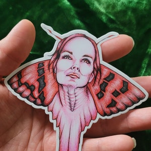 May include: A sticker featuring a woman's face with red and black butterfly wings. The woman's face is drawn in detail, with a soft, pink hue. The butterfly wings are spread out, with a black and red pattern. The sticker is signed "2019 RAINBOW".