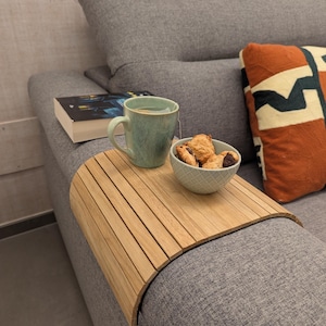 Op de afbeelding: Een houten armleuningtafel voor de bank houdt een turquoise mok, een kleine kom met koekjes en een boek vast. De tafel staat op een grijze bank, met een decoratief kussen op de achtergrond. De tafel is van licht hout.
