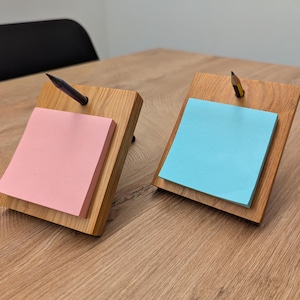 Könnte beinhalten: Zwei Holz-Notizhalter mit Bleistiften. Einer hat einen rosa Haftnotizblock, der andere einen blauen. Die Halter stehen auf einem Holztisch.