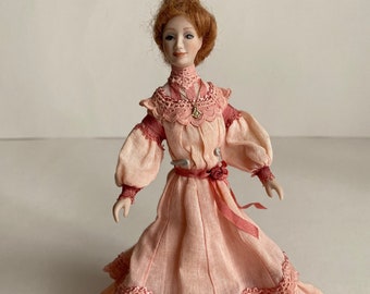 May include: A porcelain doll wearing a pink dress with lace trim and a ribbon. The doll has red hair and is wearing a necklace with a pendant.