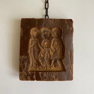 May include: A brown wax icon depicting three figures, likely a religious scene, hanging from a metal chain. The figures are embossed in the wax and have a detailed design.