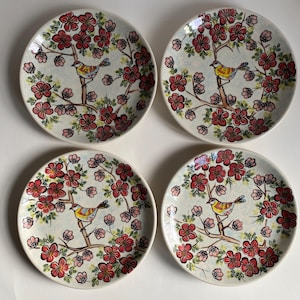 May include: Four ceramic plates with a hand-painted design. Each plate features a bird perched on a branch surrounded by red and pink flowers. The plates have a white background and are arranged in a square.