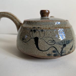 May include: A ceramic teapot with a speckled, gray-brown finish. The teapot features a curved handle and a lid with a small, brown knob. Black floral designs are painted on the body. A decorative item for the kitchen.