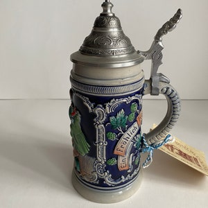 May include: A blue and white ceramic beer stein with a pewter lid and handle. The stein features a painted scene of a woman in traditional German dress and the text "Ein Frohlich".