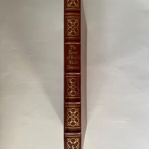 Puede incluir: Un libro de tapa dura, "The Essays of Ralph Waldo Emerson", con cubierta de cuero marr&oacute;n y detalles dorados. La columna vertebral presenta el t&iacute;tulo y patrones decorativos. Una cinta roja de marcador es visible.