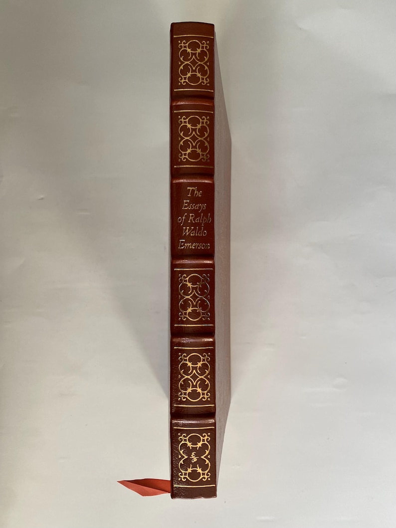 Puede incluir: Un libro de tapa dura, "The Essays of Ralph Waldo Emerson", con cubierta de cuero marr&oacute;n y detalles dorados. La columna vertebral presenta el t&iacute;tulo y patrones decorativos. Una cinta roja de marcador es visible.